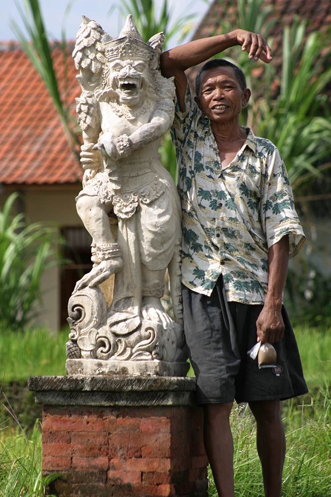 Smiling older man leaning against an elaborately carved statue and holding a small bag containing something brown.