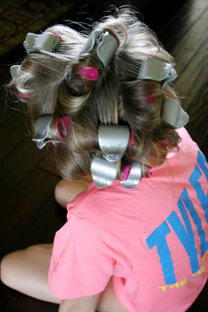 Looking down at a girl sitting cross-legged on the floor wearing a pink and blue tshirt with hot pink and silver rollers in her hair. 