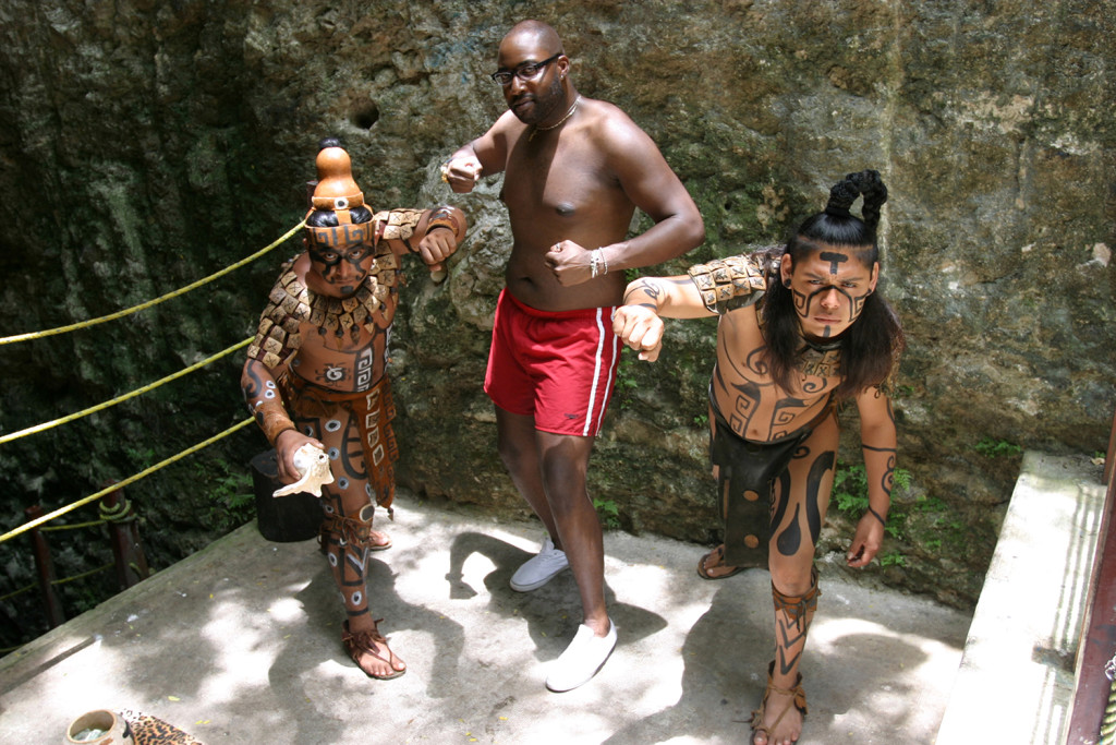 A tourist posing as a Mayan warrior in between two Mayan descendants dressed as warriors. 