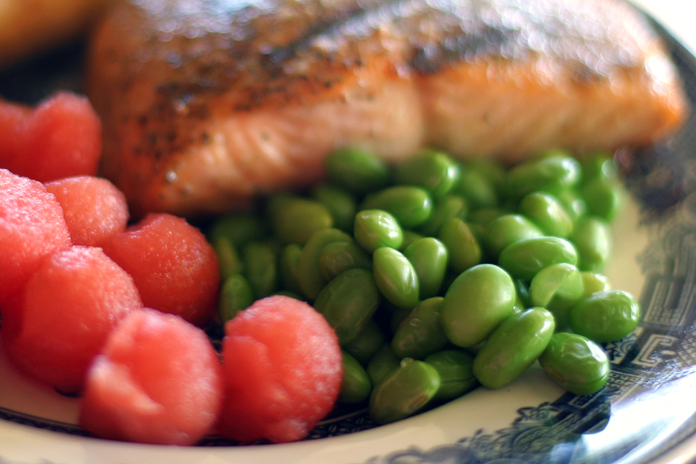A blue and white plate filled with bright green edamame, juicy red watermelon balls, and a freshly grilled slice of salmon with lots of seasoning.