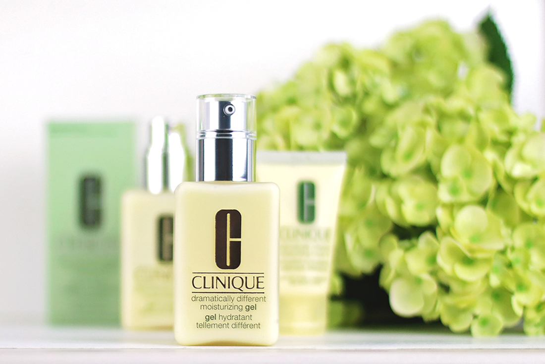 3 buttery-yellow colored bottles filled with Clinique Dramatically Different Gel lying on a table next to a box of Clinique packaging and bright green hydrangeas with a white background.