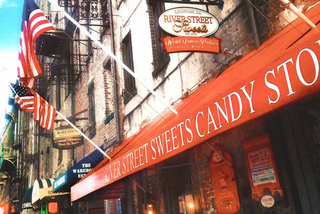 On River Street in Savannah, a storefront is lined with colorful flags and a bright red awning with white lettering that reads River Street Sweets Candy Store.