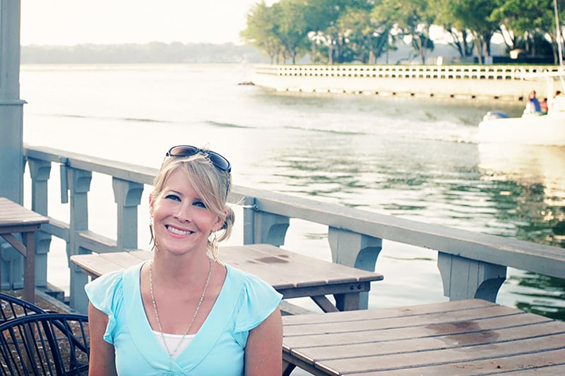 Girl in a short sleeved turquoise blue top smiling as a boat enters the marina behind her.