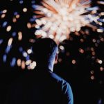 A man seen from the back watching a burst of red white and blue fireworks.