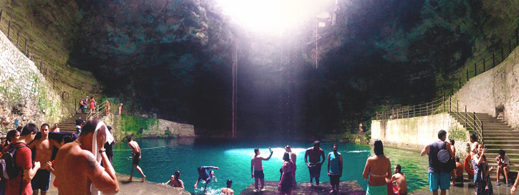 Wide angle view showing the opening to the sky above Cenote Hubiku with sunlight streaming down into a caverous opening where swimmers encircle turquoise water.