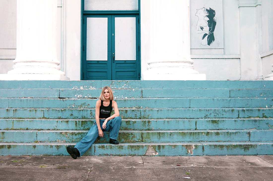Teenage girl in jeans and a black tank top sitting on worn, blue steps at a large white school building.