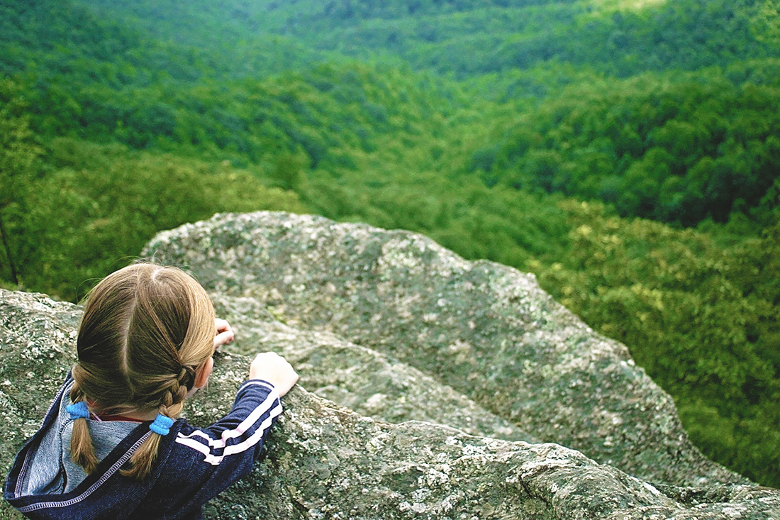A small girl peering over the edge of a steep rock to a lush green valley hundreds of feet below.