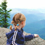 A young girl in a navy blue and pink outfit with her hair in pigtails looks over The Blowing Rock at the blue and green valley below.