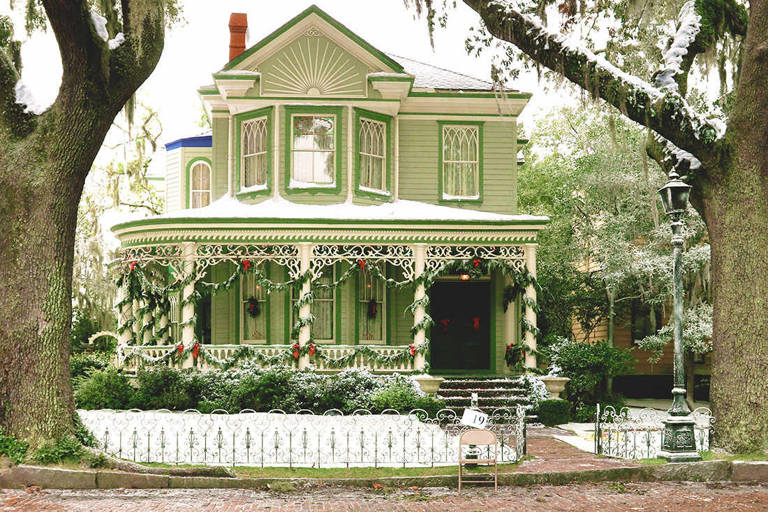 A snowy scene of a 2-story green Victorian-style home with a wraparound front porch covered in traditional Christmas wreaths and garlands.