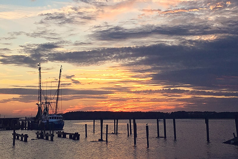 A docked shrimp boat and old dock pilings in the foreground with a purple and lavender sunset fading into yellow and orange over the horizon.
