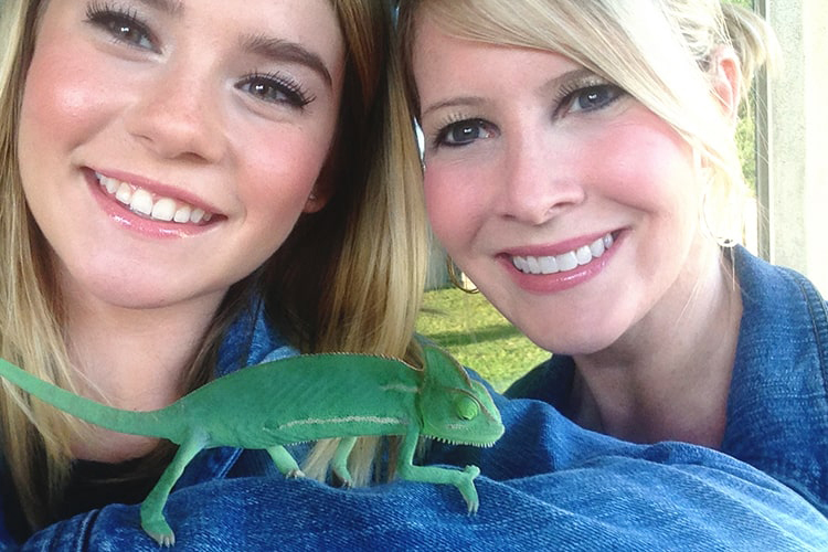 Two girls in matching denim jackets smiling as a bright green chameleon crawls on one girl's arm.