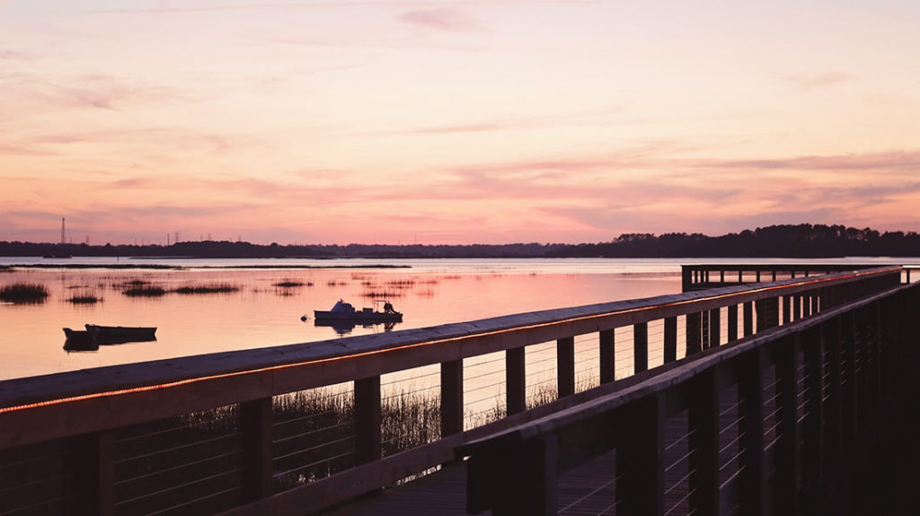 Silhouetted boats in the water with a peachy-lavender sunset as seen from a wooden pier on Hilton Head Island.