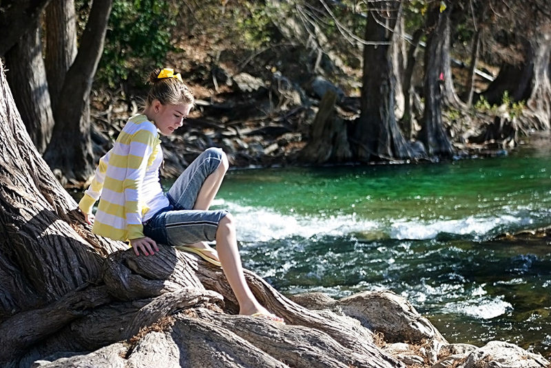 A cute young girl with a big yellow bow in her hair is about to stick her toes in the Guadalupe River. 